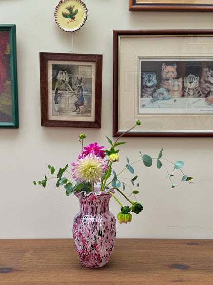 Pink vase with flowers on a wooden table against a wall with framed pictures